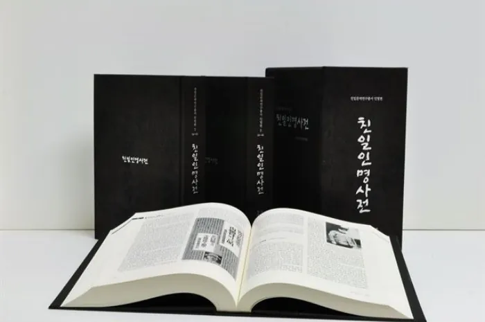검은 표지의 책 여러 권이 세워져 있고 그 앞에 한 권이 펼쳐져 있는 친일인명사전.